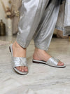 A person wearing shiny silver slide sandals with a bow detail on the front, paired with matching silver pants, standing on a light marble floor.